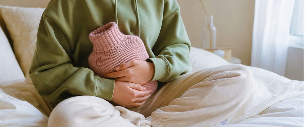 Person sitzt im Schneidersitz auf einem Bett und hält eine rosa Wärmflasche mit beiden Händen vor dem Bauch, trägt grünen Hoodie und helle Jogginghose