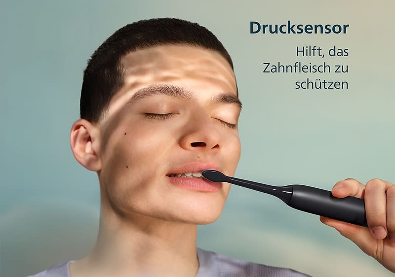 Eine Person hält eine schwarze elektrische Zahnbürste. Der Text „Drucksensor hilft, das Zahnfleisch zu schützen“ ist oben rechts im Bild zu sehen.