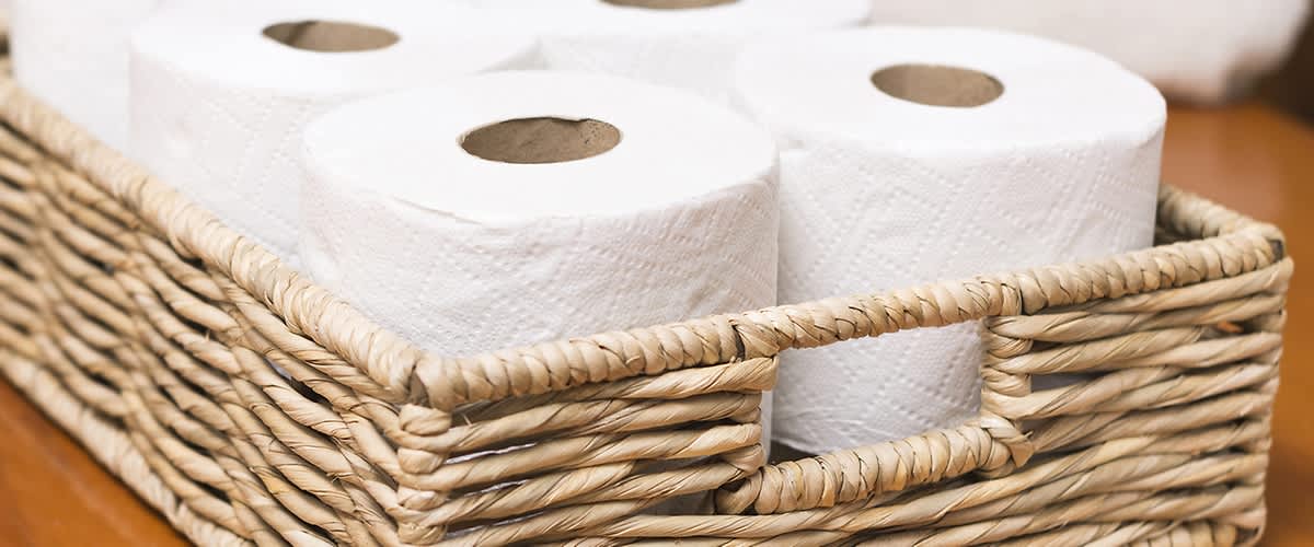 Vier weiße Toilettenpapierrollen in einem geflochtenen Korb auf einer Holzoberfläche