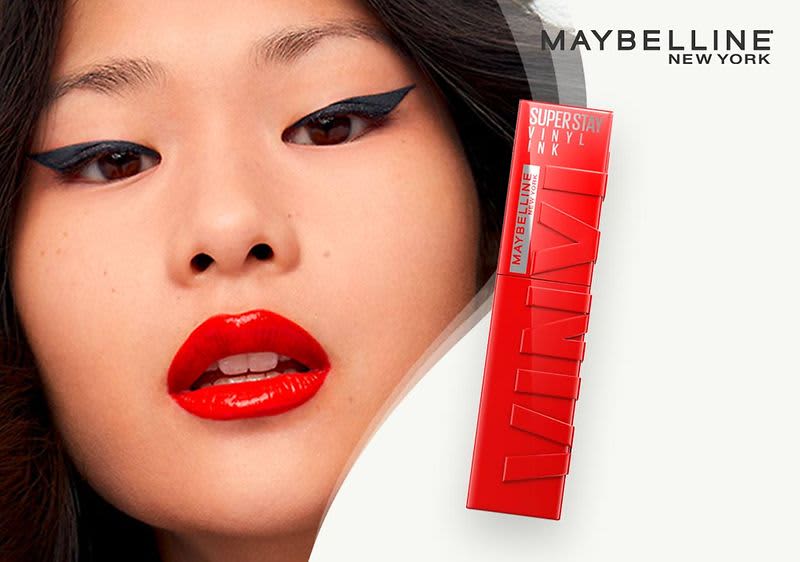 Eine rote Verpackung mit der Aufschrift SUPER STAY MATTE INK in weißer Schrift und darunter MAYBELLINE NEW YORK in schwarzer Schrift, die Verpackung ist länglich und rechteckig. Der Hintergrund ist hell und ein Teil eines Gesichts ist dargestellt.