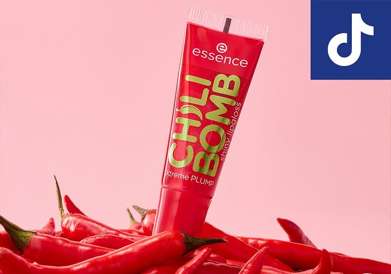 Eine rote Tube mit der Aufschrift essence CHILI BOMB extreme PLUMP instant lipgloss steht auf einer Ansammlung von roten Chilischoten vor einem rosa Hintergrund, oben rechts ist ein blaues Quadrat mit einem weißen TikTok-Logo zu sehen