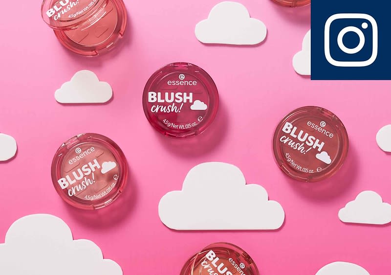 Mehrere runde Blush Dosen mit der Aufschrift „Essence Blush Crush!“ liegen auf einem rosa Hintergrund, umgeben von weißen Wolken aus Papier