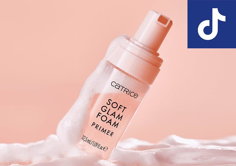 Eine rosafarbene Flasche mit einem Pumpspender, auf der in schwarzer Schrift CATRICE SOFT GLAM FOAM PRIMER 45 ml/1.01 fl.oz steht, ist von weißem Schaum umgeben, im Hintergrund ist ein hellrosa Farbton zu sehen, oben rechts befindet sich ein blaues Quadrat mit einem weißen TikTok-Logo.