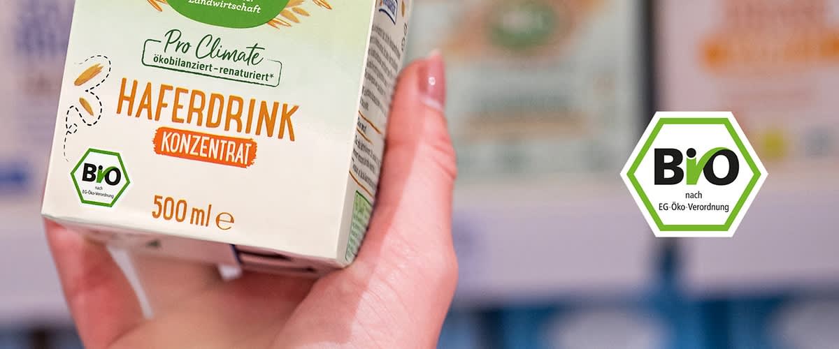 Eine Hand hält eine Verpackung mit der Aufschrift „Haferdrink Konzentrat“ und „500 ml“. Oben steht „Pro Climate Ökobilanziert – Renaturiert“. Ein Bio-Siegel ist sichtbar.