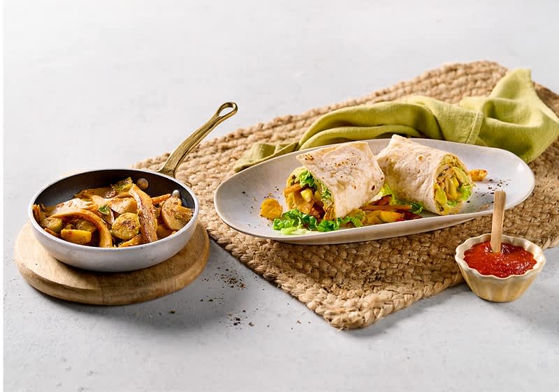 Ein Teller mit zwei halbierten Wraps, gefüllt mit Gemüse und Fleisch, daneben eine Pfanne mit gebratenem Fleisch und Gemüse sowie ein kleines Schälchen mit roter Soße auf einem geflochtenen Tischset.