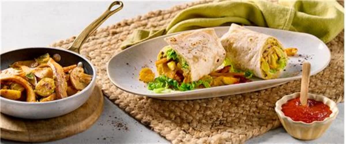 Ein Teller mit zwei halbierten Wraps, gefüllt mit Gemüse und Fleisch, daneben eine Pfanne mit gebratenem Fleisch und Gemüse sowie ein kleines Schälchen mit roter Soße auf einem geflochtenen Tischset.