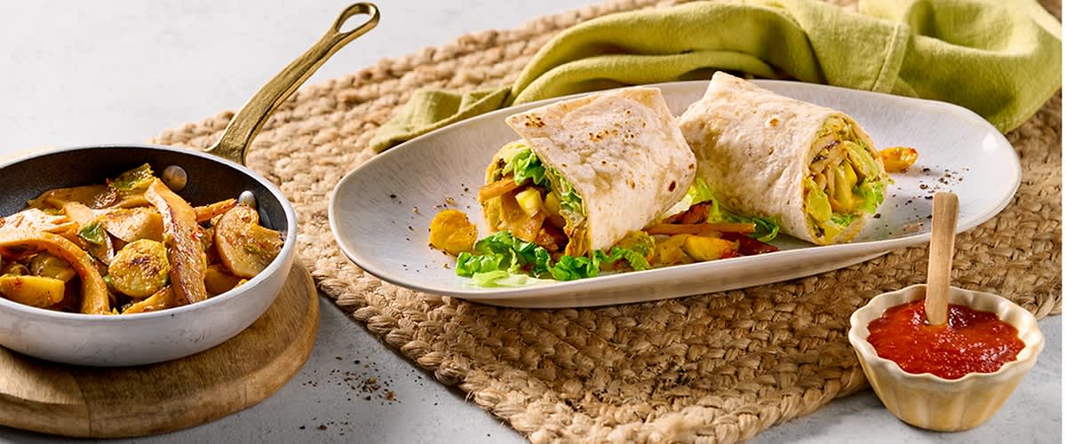 Ein Teller mit zwei halbierten Wraps, gefüllt mit Gemüse und Fleisch, daneben eine Pfanne mit gebratenem Fleisch und Gemüse sowie ein kleines Schälchen mit roter Soße auf einem geflochtenen Tischset.