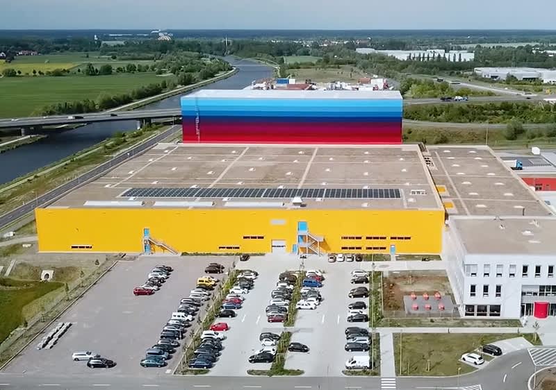 VZ Wustermark: Ein großes, modernes Gebäude mit einer gelben Fassade und einem farbenfrohen Streifenmuster auf der oberen Hälfte steht in einer weitläufigen Umgebung, im Vordergrund sind zahlreiche geparkte Autos auf einem Parkplatz zu sehen, im Hintergrund erstreckt sich eine grüne Landschaft mit einem Fluss und einer Brücke