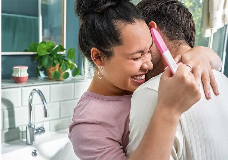 Eine Frau mit dunklem, hochgestecktem Haar und großen Ohrringen umarmt einen Mann und hält einen Schwangerschaftstest in der Hand, während im Hintergrund eine grüne Pflanze und ein Waschbecken mit Wasserhahn zu sehen sind