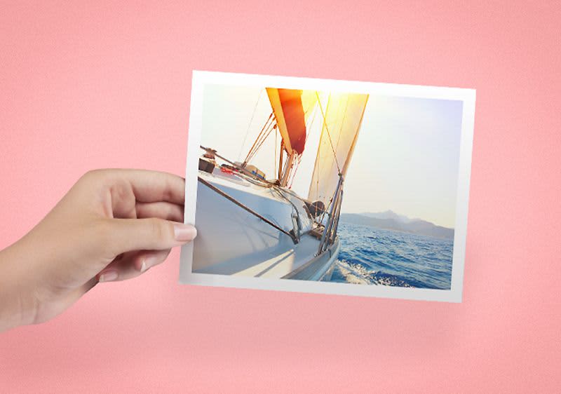 Eine Hand hält ein Foto von einem Segelboot auf dem Meer, das von Sonnenlicht beleuchtet wird. Der Hintergrund ist rosa.