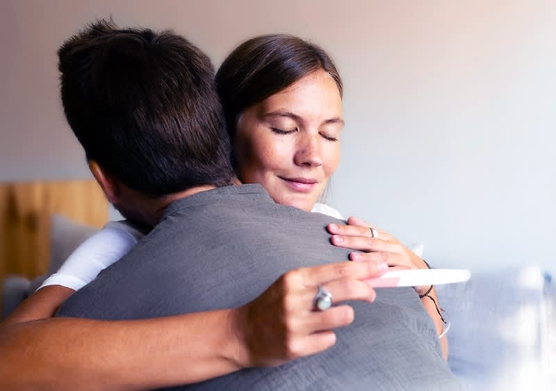 Ein Mann mit dunklem Haar und grauem T-Shirt umarmt eine Person, deren Gesicht unscharf ist, während er einen Schwangerschaftstest in der Hand hält, der ein positives Ergebnis anzeigt, die Szene vermittelt eine emotionale und freudige Stimmung