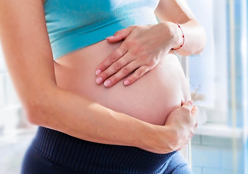 Eine schwangere Frau steht in einem Badezimmer und hält ihren Bauch mit beiden Händen, sie trägt ein hellblaues Sporttop und eine schwarze Leggings, im Hintergrund sind ein Waschbecken, ein Spiegel und eine Pflanze auf einem Fensterbrett zu sehen