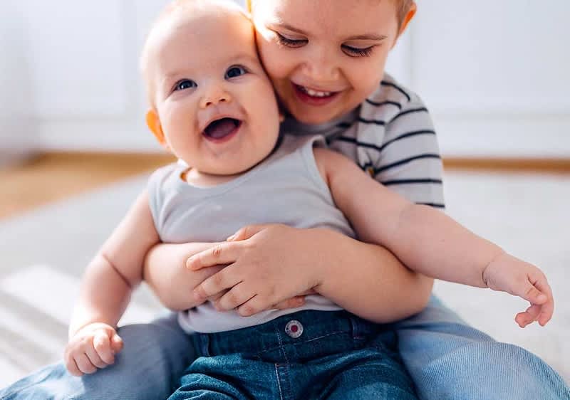 Ein Kleinkind in einem grauen ärmellosen Oberteil und blauer Jeans sitzt auf dem Boden und wird von einem älteren Kind in einem gestreiften T-Shirt liebevoll umarmt, beide Gesichter sind unscharf gemacht.