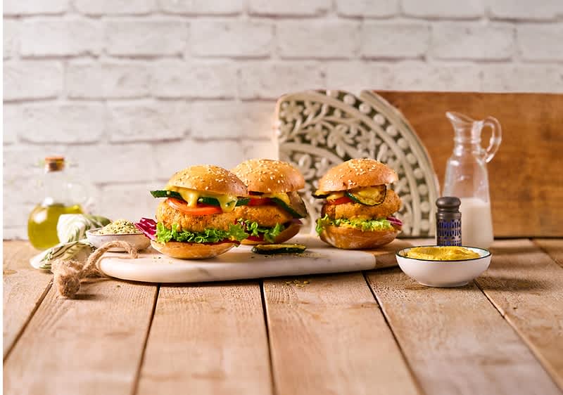 Drei vegetarische Burger mit Sesambrötchen, Salat, Tomaten und Gurken auf einem weißen Tablett, daneben eine Karaffe mit Milch und eine kleine Schale mit gelber Sauce auf einem Holztisch