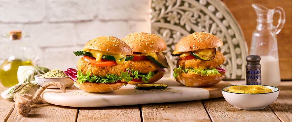 Drei vegetarische Burger mit Sesambrötchen, Salat, Tomaten und Gurken auf einem weißen Tablett, daneben eine Karaffe mit Milch und eine kleine Schale mit gelber Sauce auf einem Holztisch