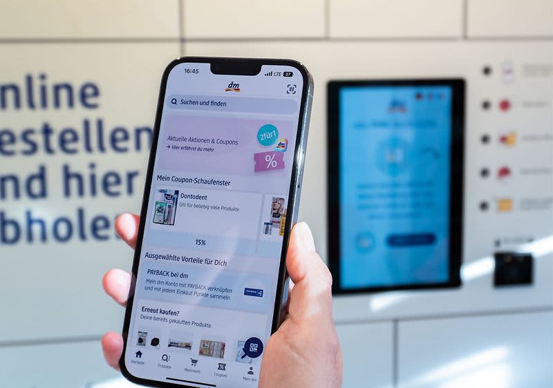Bild eines Smartphones mit laufender Mein dm-App vor eine Abholstation in einem dm-Markt