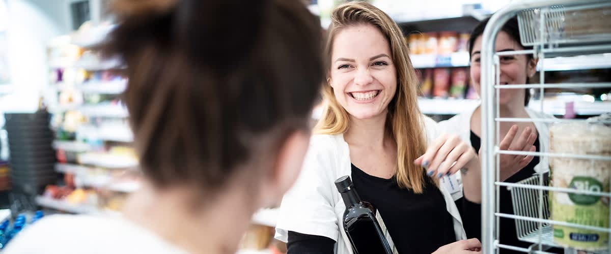 Eine dm-Mitarbeiterin mit langen blonden Haaren trägt eine schwarze Flasche unter ihrem Arm und steht neben einem Einkaufswagen, während im Hintergrund Regale mit verschiedenen Produkten zu sehen sind