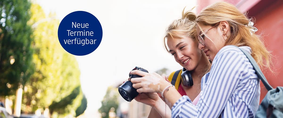 Zwei junge Frauen betrachten gemeinsam eine digitale Kamera, eine zeigt etwas auf dem Kameramonitor, beide lächeln leicht, links oben ein blauer Kreis mit weißer Schrift „Neue Termine verfügbar“