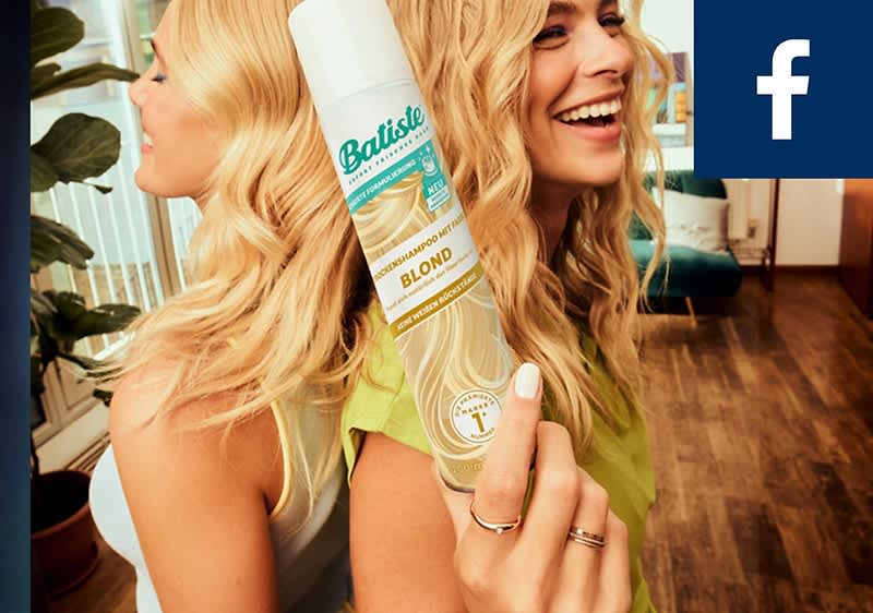 Zwei blond gelockte Frauen Rücken an Rücken, vorn hält eine Hand ein weißes Batiste Trockenshampoo-Spray mit Aufschrift „Blond“, im Bild oben rechts kleines blaues Facebook-Symbol