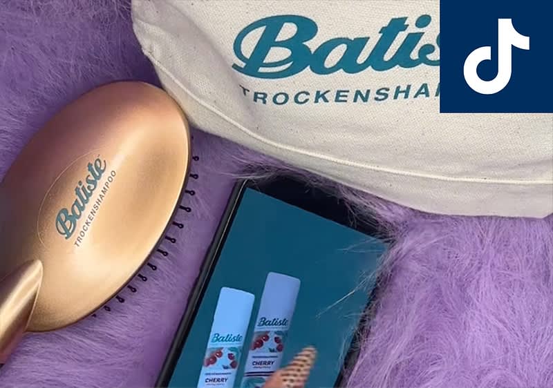 Eine goldene Haarbürste mit der Aufschrift „Batiste Trockenshampoo“ liegt auf einem flauschigen, lila Untergrund neben einer Tasche mit dem Schriftzug „Batiste Trockenshampoo“ und einem Smartphone, das zwei Batiste Trockenshampoo-Produkte zeigt. Oben rechts ist das TikTok-Logo zu sehen