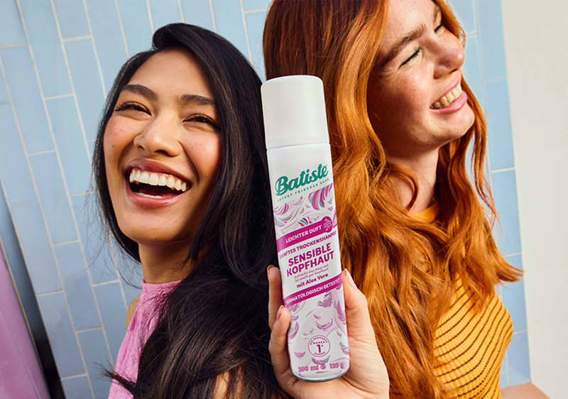 Zwei lachende Frauen stehen nebeneinander, vorne eine dunkelhaarige Frau, hinten eine rothaarige Frau, eine Hand hält eine weiße Batiste-Trockenshampoo-Dose mit lila Aufschrift „Sensible Kopfhaut“ sichtbar in Bildmitte
