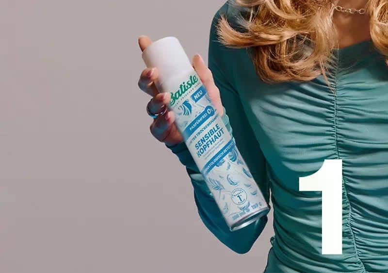 Frau in türkisfarbenem, gerafftem Oberteil hält eine weiße und türkis gestaltete Spraydose mit Markenname „Batiste“ und Aufschrift „Sensitive Kopfhaut“ sichtbar, rechts große weiße Ziffer „1“