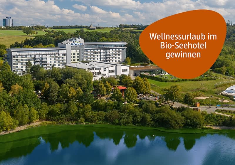 Großes Hotelgebäude am Ufer eines Sees, rechts orangefarbene element mit dem Text „Wellnessurlaub im Bio-Seehotel gewinnen“