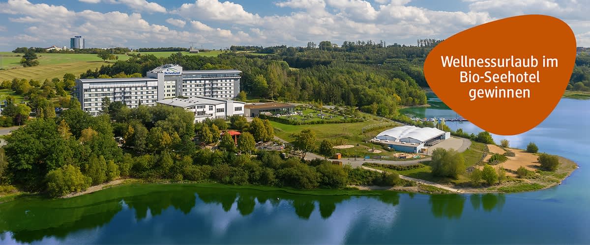 Großes Hotelgebäude am Ufer eines Sees, rechts orangefarbene element mit dem Text „Wellnessurlaub im Bio-Seehotel gewinnen“