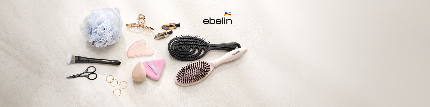 Verschiedene Haar- und Make-up-Accessoires auf hellem Untergrund: zwei Haarbürsten (eine schwarz, geschwungene Entwirrbürste, eine rosafarbene Bürste mit Metallstiften), zwei beige Make-up-Schwämmchen und ein rosafarbenes, dreieckiges Schwämmchen mit Aufdruck „ebelin“, zwei rosa Gua-Sha-Steine, zwei tortoisefarbene Haarklammern, eine goldfarbene Haarspange sowie vier goldene Ringe, oben rechts Schriftzug „ebelin“ und kleines dm-Logo
