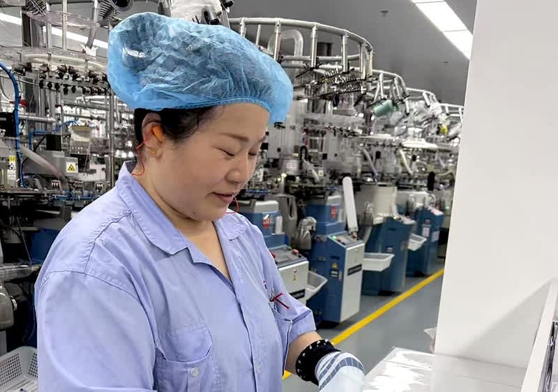 Arbeiterin in blauer Schutzkleidung und Haube steht an einer Maschine in einer Produktionshalle mit Reihen von Industrieanlagen im Hintergrund