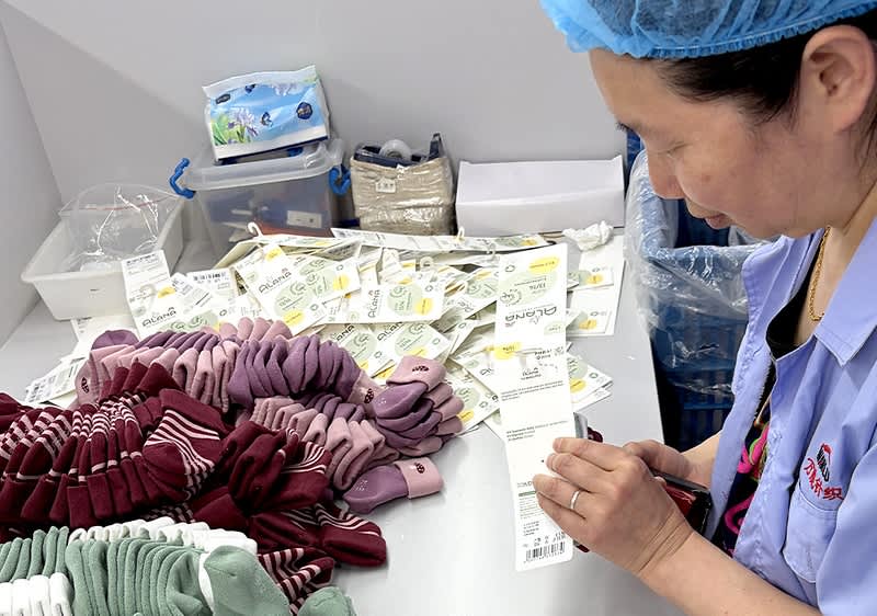 Frau mit Haarnetz und Dienstkleidung schneidet an einem Tisch aus einem Stapel Etiketten einzelne Preisschilder, auf dem Tisch liegen gestapelte Socken in verschiedenen Farben sowie Verpackungsmaterial und Kunststoffboxen