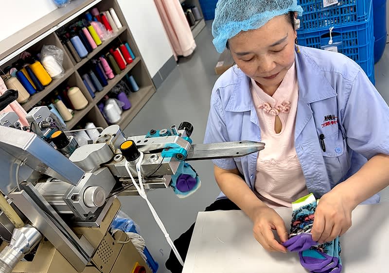 Frau an einer Nähmaschine faltet bunte Socken, trägt Arbeitskleidung und Haarnetz, Regal mit Garnspulen im Hintergrund