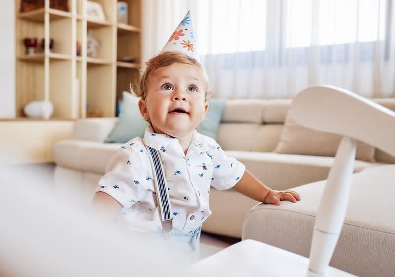 Kleines Kleinkind mit Papierspitzenpartyhut und hellblauem Hemd mit Hosenträgern sitzt an einem Sofa im Wohnzimmer und stützt sich mit einer Hand auf die Armlehne, im Hintergrund Regal und Kissen sichtbar