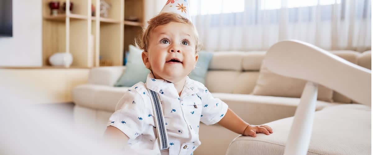 Kleines Kleinkind mit Papierspitzenpartyhut und hellblauem Hemd mit Hosenträgern sitzt an einem Sofa im Wohnzimmer und stützt sich mit einer Hand auf die Armlehne, im Hintergrund Regal und Kissen sichtbar