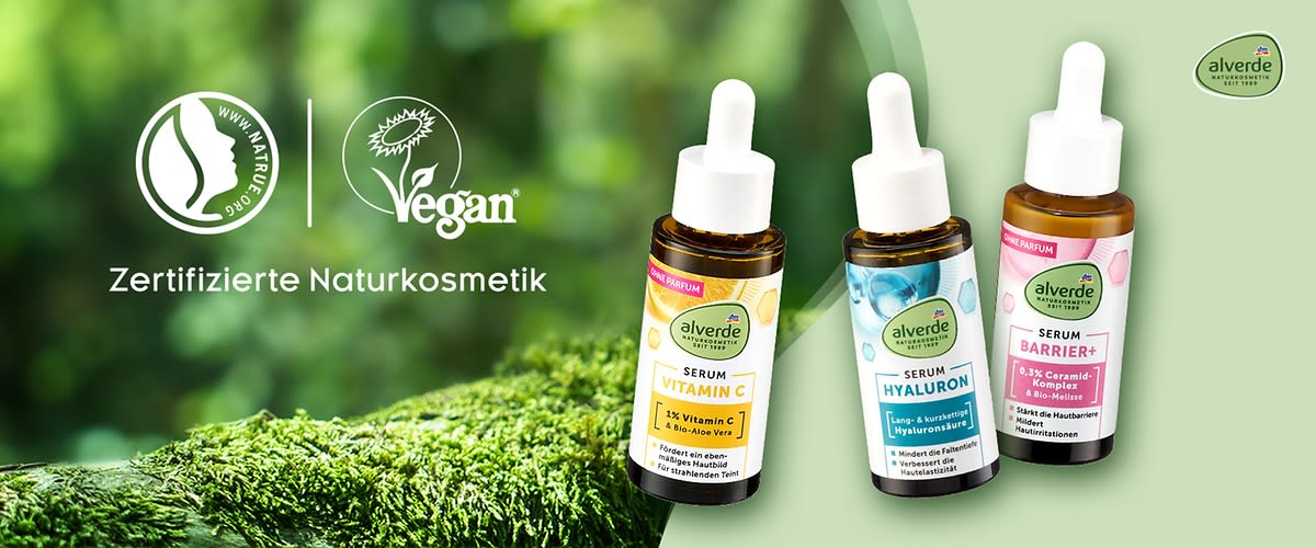 Drei Flaschen Alverde-Serum mit Pipetten vor einem grünen Hintergrund. Links sind die Logos für zertifizierte Naturkosmetik und vegan zu sehen. Die Flaschen sind mit den Aufschriften „Vitamin C“, „Hyaluron“ und „Barrier+“ versehen. Oben rechts befindet sich das Alverde-Logo.