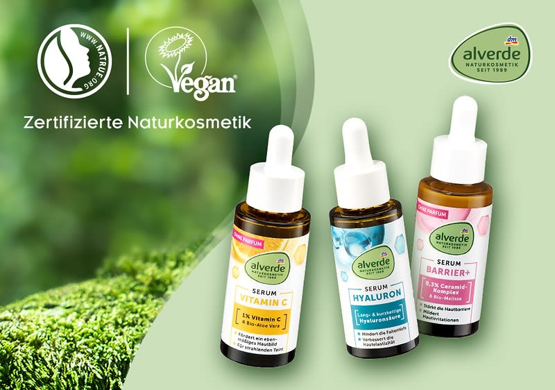 Drei Flaschen Alverde-Serum mit Pipette, jeweils mit unterschiedlicher Aufschrift: Vitamin C, Hyaluron und Barrier. Links oben Logos für zertifizierte Naturkosmetik und vegan. Rechts oben das Alverde-Logo. Im Hintergrund unscharfes Grün.
