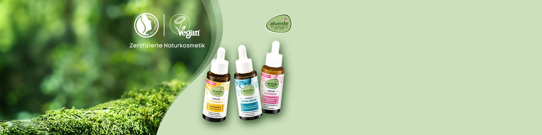 Drei Flaschen Alverde-Serum mit Pipette, jeweils mit unterschiedlicher Aufschrift: Vitamin C, Hyaluron und Barrier. Links oben Logos für zertifizierte Naturkosmetik und vegan. Rechts oben das Alverde-Logo. Im Hintergrund unscharfes Grün.