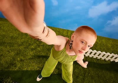 Eine Person in einem grünen Outfit steht auf einer grünen Wiese und streckt ihren Arm in die Kamera, im Hintergrund ein weißer Zaun und blauer Himmel mit Wolken