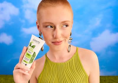 Eine Person in einem grünen Oberteil hält eine Tube mit der Aufschrift „SANTE Family Bio Aloe Vera Feuchtigkeitspflege“ vor einem blauen Himmel mit Wolken