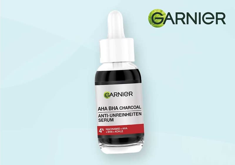 Garnier AHA BHA Charcoal Anti-Unreinheiten Serum in klarer Pipettenflasche mit schwarzem Inhalt und weißer Etikette, auf der „AHA BHA CHARCOAL ANTI-UNREINHEITEN SERUM“ und „4% NIACINAMID + AHA + BHA + KOHLE“ zu lesen sind, vor hellem Hintergrund und Garnier-Logo oben rechts