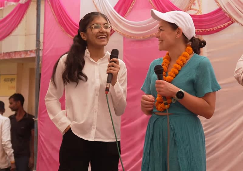 Zwei Personen stehen nebeneinander und halten Mikrofone. Die Person links trägt ein weißes Hemd und eine schwarze Hose, die Person rechts trägt ein blaues Kleid, eine weiße Kappe und eine Blumenkette. Im Hintergrund sind rosa und weiße Stoffbahnen zu sehen.