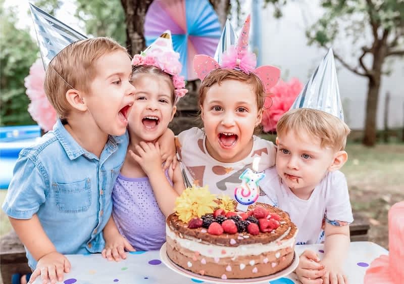 Vier Kinder mit Partyhüten stehen eng beieinander hinter einem Geburtstagskuchen auf einem bunt gepunkteten Tisch, auf dem Kuchen liegen Beeren, Blumen und eine Kerze in Form der Zahl 3, im Hintergrund Gartenparty-Dekoration mit Papierfächern und Luftballons