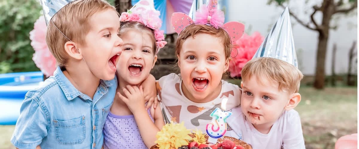 Vier Kinder mit Partyhüten stehen eng beieinander hinter einem Geburtstagskuchen auf einem bunt gepunkteten Tisch, auf dem Kuchen liegen Beeren, Blumen und eine Kerze in Form der Zahl 3, im Hintergrund Gartenparty-Dekoration mit Papierfächern und Luftballons