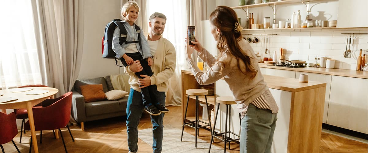 Ein Mann hält ein Kind mit Schulrucksack auf dem Arm, während eine Frau ein Foto von ihnen in einer modernen Küche macht