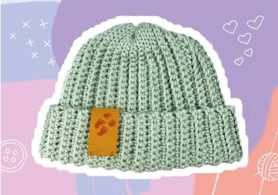 Eine hellgrüne Strickmütze mit einem umgeschlagenen Rand, auf dem ein braunes Etikett mit roten Fußabdrücken und Handabdrücken angebracht ist, vor einem Hintergrund aus lila und rosa Farbflächen mit weißen Herzen, Kreuzen und einem Knopf-Symbol