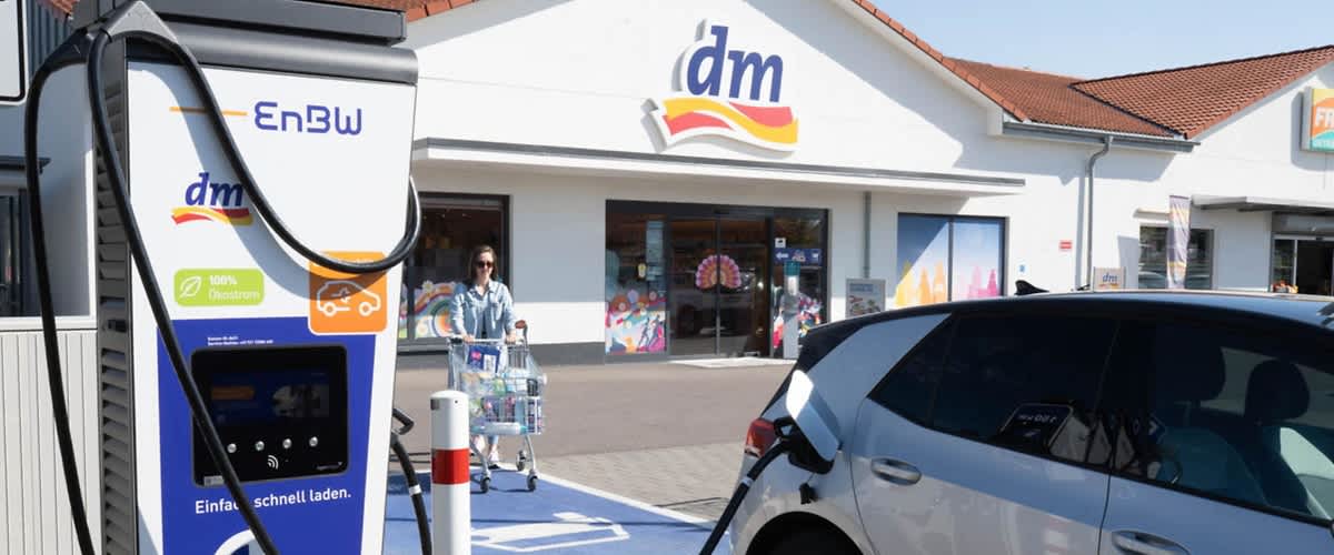 Eine Frau schiebt einen Einkaufswagen auf dem Parkplatz eines Drogeriemarktes der Kette dm, während ein Elektroauto an einer EnBW-Ladestation aufgeladen wird, im Hintergrund ist das Gebäude mit dem dm-Logo zu sehen