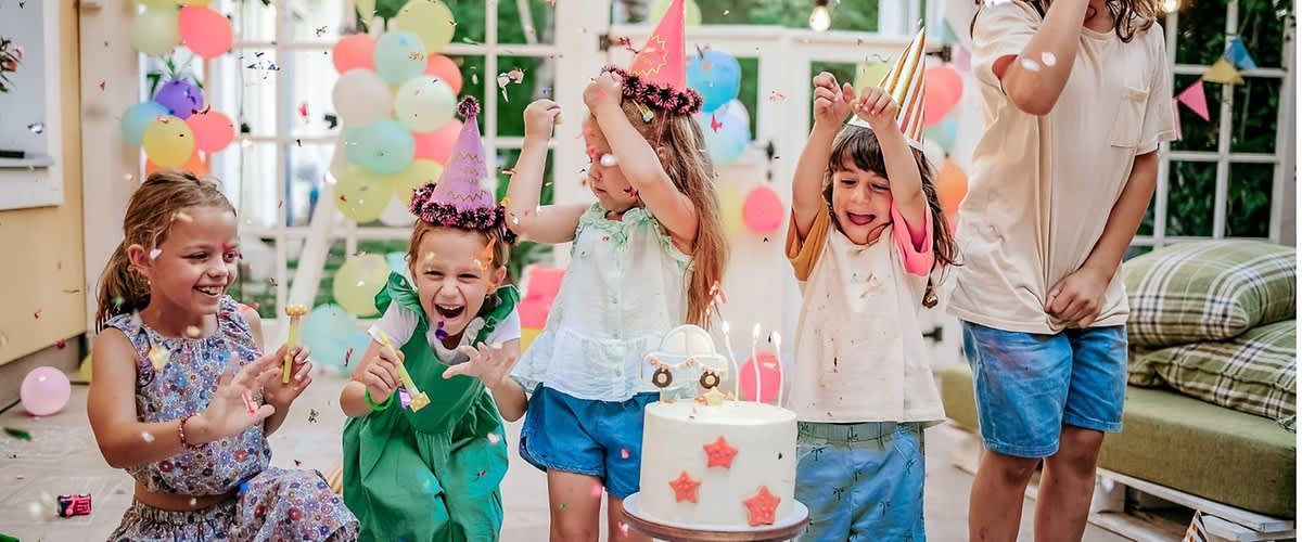 Kinder feiern Geburtstagsfeier in einem hellen Raum, fünf Kinder tragen bunte Partyhüte und stehen hinter einem zweistöckigen Geburtstagskuchen mit roten Sternen auf einer Holzständer, im Hintergrund bunte Luftballons und Wimpelgirlanden, einige Kinder werfen Konfetti und wirken fröhlich