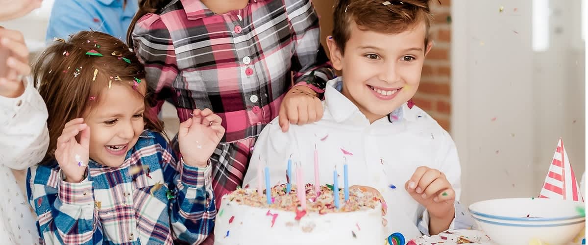 Vier Kinder um einen Geburtstagskuchen auf einem Tisch, auf dem bunte Konfetti liegt; der runde Kuchen ist mit weißer Glasur, bunten Streuseln und fünf brennenden Kerzen dekoriert, ein Kind trägt ein rot-kariertes Hemd und ein anderes ein weißes Hemd, im Hintergrund eine Ziegelwand und ein Fenster ohne weitere Details