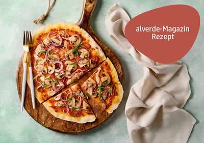 Auf einem runden Holzbrett liegt ei Teig mit Tomatensauce, Pilzen, roten Zwiebeln und grünen Kräutern, daneben ein Messer und eine Gabel, auf einem hellgrünen Untergrund mit einem beigen Stofftuch, oben rechts steht in einem roten Feld „alverde-Magazin Rezept“
