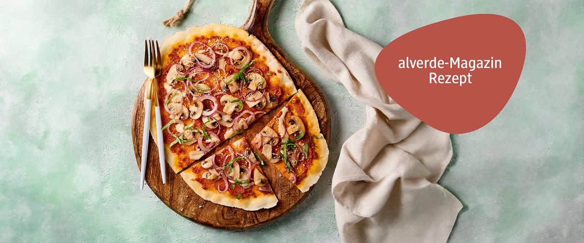 Auf einem runden Holzbrett liegt ei Teig  mit Tomatensauce, Pilzen, roten Zwiebeln und grünen Kräutern, daneben ein Messer und eine Gabel, auf einem hellgrünen Untergrund mit einem beigen Stofftuch, oben rechts steht in einem roten Feld „alverde-Magazin Rezept“